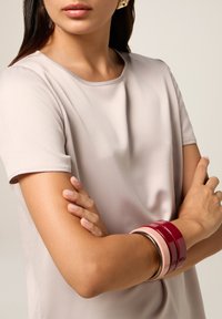 Top a maniche corte di un beige chiaro con una superficie liscia. Polso mostrato con bracciali lucidi impilati in rosso porpora, rosa chiaro e argento.