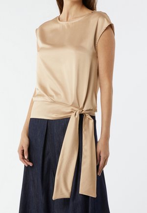 Top sin mangas de satén beige con detalle de lazo lateral, usado con falda plisada oscura, mostrado desde la parte superior del torso hasta la mitad del muslo.