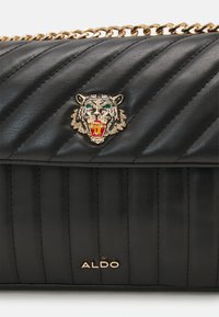 Sac à main en simili cuir noir matelassé avec une chaîne dorée, un ornement en forme de tête de tigre et un logo en relief en bas.
