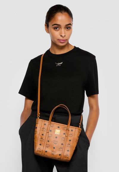 Woman in black shirt and dark pants carrying a tan patterned leather handbag with shoulder strap and handles against a plain background.