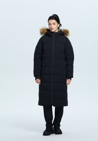 Schwarze Daunenjacke mit langem Schnitt, rechteckigem Steppmuster, Reißverschluss und Kapuze mit Kunstpelz. Getragen über dunklen Hosen und Stiefeln.