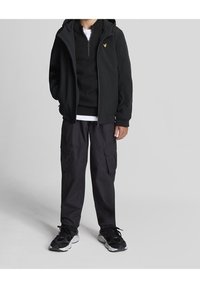 Veste zippée noire avec capuche, superposée sur un pull noir et une chemise blanche. Pantalons cargo foncés avec poches, associés à des baskets noires.