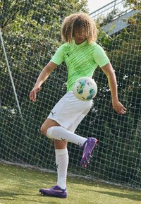 Ljusgrön t-shirt med kort ärm och mönster, vita shorts, lila fotbollsskor och knälånga vita strumpor. Fotboll i rörelse.