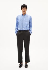 Blauw-wit gestreepte overhemd met kraag, gecombineerd met aansluitende donkergrijze broeken en zwarte instappers, staande met de handen in de zakken.