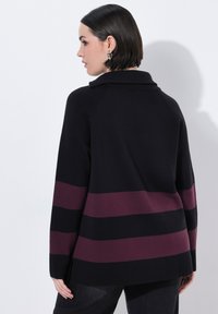 Kvinde med kort mørkt hår, iført en sort sweater med to brede horisontale mørkeblå striber på ryggen og ærmerne.