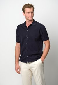 Camicia button-up a maniche corte di colore blu navy con tessuto a trama, dotata di colletto e taschino sul petto, abbinata a pantaloni color crema.
