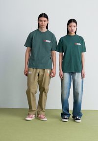 Tricouri din bumbac verde cu logo, asortate cu pantaloni bej cu branding NBA. Femeia poartă blugi de un albastru deschis și adidași negri.