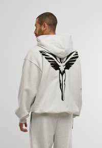 Hombre de perfil vistiendo una sudadera gris clara y pantalones de chándal, con una gran figura alada estilizada negra en la parte posterior de la sudadera.