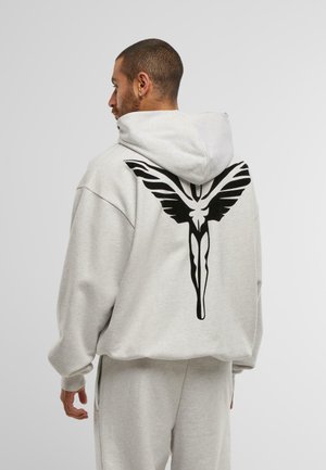 Mann, der seitlich steht und einen hellgrauen Kapuzenpullover sowie Jogginghosen trägt, mit einer großen schwarzen stilisierten geflügelten Figur auf dem Rücken des Pullovers.