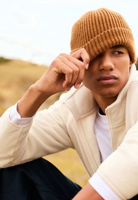 Braune gerippte Strickmütze, tief auf dem Kopf getragen, cremefarbene Fleece-Jacke mit Reißverschluss über weißem Hemd. Die Person stützt die Hand an das Gesicht, der Hintergrund ist verschwommen.