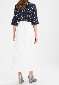 Mujer de pie con una blusa de lunares en azul marino, una falda midi blanca y zapatos de tacón bloque plateados, vista desde la parte trasera.