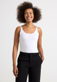 Hvid ærmeløs bodysuit med en glat tekstur, ledsaget af sorte skræddersyede bukser. Outfittet har en rund halsudskæring og slim fit.