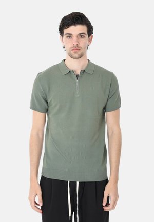 Giovane uomo visto frontalmente che indossa una polo a maniche corte in maglia verde oliva con una zip frontale e pantaloni neri con coulisse bianche.