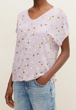 Kobieta ubrana w liliowy, krótki rękaw, dekolt w kształcie litery V, z małym, rozmieszczonym żółto-białym kwiatowym nadrukiem, połączony z jasnoniebieskimi dżinsami.