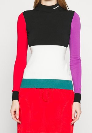 Femme portant un haut à manches longues color block avec des sections noires, blanches, turquoise, rouges et violettes, associé à une jupe rouge.