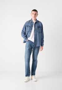 Denimjack met knoopsluiting, borstzakken en een relaxte pasvorm, gecombineerd met lichtblauwe jeans en witte sneakers. Effen wit t-shirt eronder.