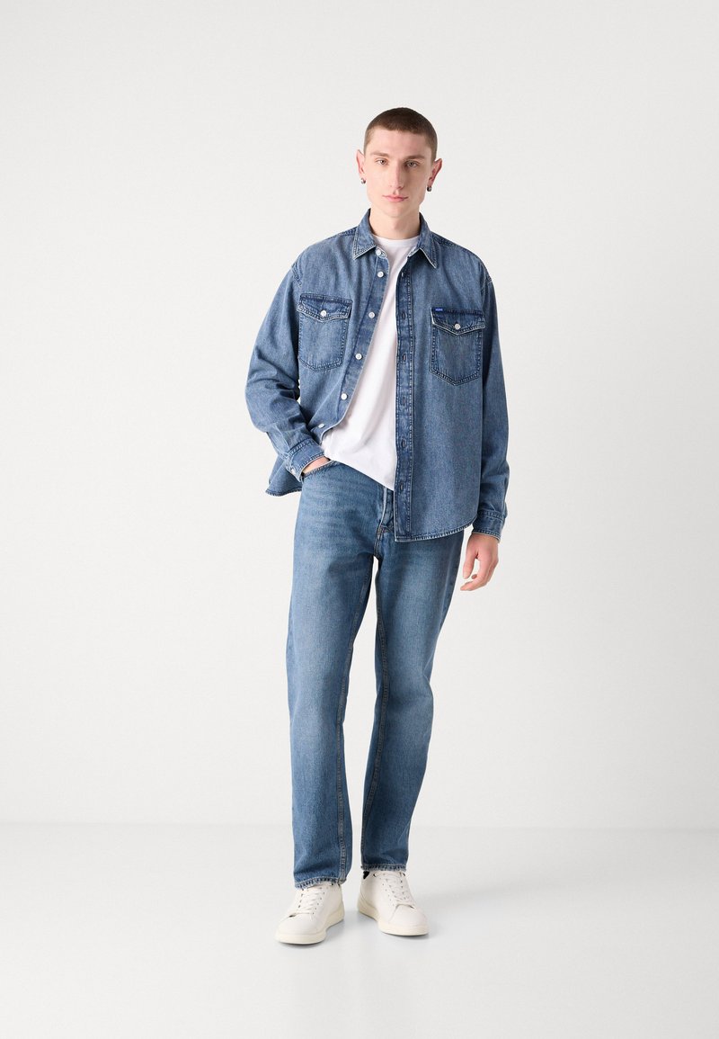 Denimjacke mit Knopfverschluss, Brusttaschen und lockerer Passform, kombiniert mit hellblauen Jeans und weißen Sneakers. Darunter ein schlichtes weißes T-Shirt.