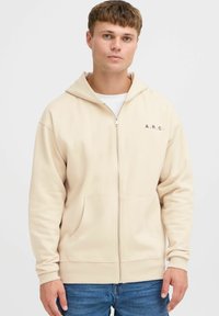Beige Kapuzenjacke mit Reißverschluss aus weichem Material, mit einer Kängurutasche, gerippten Bündchen und einem kleinen aufgedruckten Logo auf der Brust.