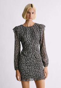 Vestido a rayas en blanco y negro con un cuerpo ceñido, mangas largas translúcidas y acentos de hombro con volantes. Tejido texturizado y detalles fruncidos.