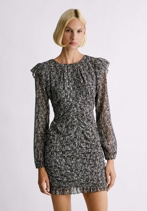 Robe à motifs noir et blanc avec un corps ajusté, des manches longues en tissu transparent et des accents de ruffles aux épaules. Tissu texturé et détails froncés.