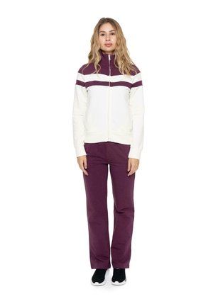 Una mujer luce una chaqueta blanca con cremallera y rayas color ciruela, junto con unos pantalones a juego del mismo color, sobre un fondo blanco liso.