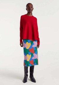 Mujer vistiendo un suéter rojo brillante, una falda de patrón de corazones coloridos y botas negras hasta la rodilla, de pie contra un fondo claro y liso.