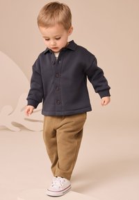 Sötét tengerészkék, gombos dzseki, alatta krémszínű csíkos ing, világosbarna nadrág és fehér tornacipő. Puha anyag, laza szabás és bordázott mandzsetták.