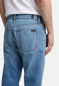 Ljusblå denimjeans med hög midja, med två bakfickor och en svart märkeetikett på midjebandet. Slät textur, avslappnad passform.