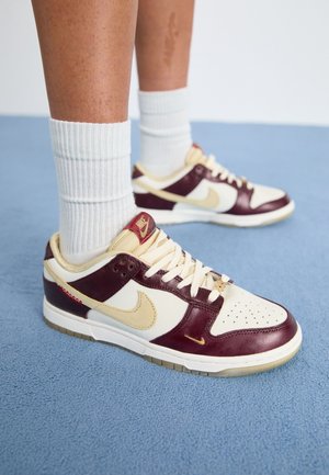 Personne portant des chaussettes blanches côtelées et des baskets Nike crème et bordeaux, debout sur un sol recouvert d'un tapis bleu.