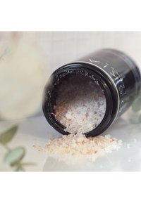 Bocal cylindrique noir avec des lettres blanches, contenant des grains de sel de teinte rose éparpillés sur une surface blanche. Feuilles vertes en arrière-plan.