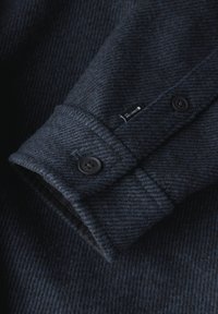 Mörk marinblå ullskjorta med en texturerad yta, försedd med en knappmanschett och en liten etikett på den inre sömmen. Dubbelknäppning.