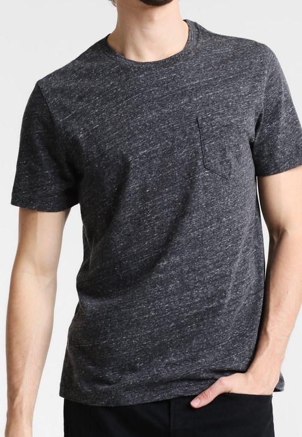 Homme portant un t-shirt gris foncé chiné à manches courtes avec une petite poche sur la poitrine, main reposant dans la poche d’un pantalon noir.