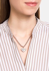 Collana in argento con pendente a forma di cuore dalla superficie liscia e bordato di piccoli strass, indossata sopra una maglietta a righe.