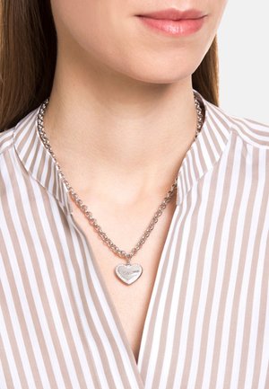 Collar de cadena de plata con un colgante en forma de corazón que tiene una superficie lisa y un borde de pequeñas incrustaciones de rhinestones, llevado sobre una camiseta a rayas.