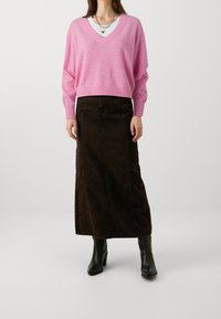 Kvinna klädd i en rosa V-ringad tröja, vit topp under, mörkbrun lång kjol, svarta klackstövlar och lager av halsband, stående framför en vit bakgrund.