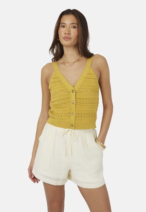 Vrouw draagt een mosterdgeel gebreid mouwloos hemd met knoopjes en witte shorts met trekkoord, staand met één hand in haar zak.