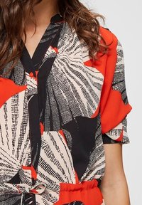 Blusa con motivo rosso e nero, con maniche corte, colletto e cintura annodata in vita. Il tessuto ha una texture morbida con disegni audaci.