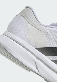 Fehér sportcipő lélegző hálós felsőrésszel, texturált részletekkel és könnyű talppal. Szürke díszítések a sarokrészen és a logón.