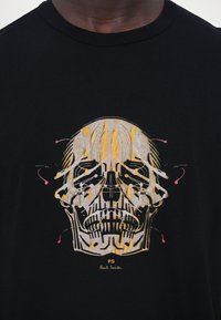 Čierne bavlnené tričko s grafickým potlačou lebky so žltými a ružovými akcentmi a textom "PS Paul Smith" na spodnej strane.