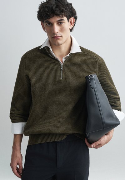 Maglione a coste verde oliva con colletto a mezza zip, abbinato a una camicia bianca con colletto. Il modello tiene una borsa nera in pelle tessuta.