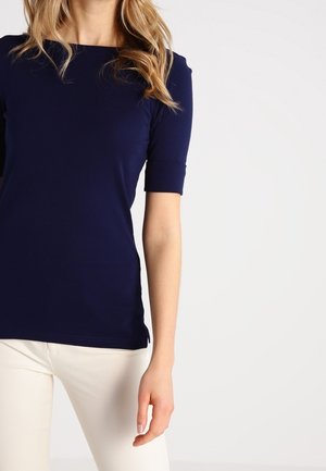 Vrouw die een nauwsluitend marineblauw T-shirt met korte mouwen en bootnek draagt, gecombineerd met gebroken witte broek, staand tegen een egaal lichte achtergrond.