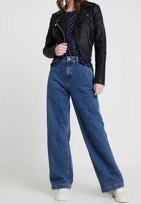 Giacca da motociclista in pelle nera sopra una blusa a motivo blu navy, abbinata a jeans in denim blu a vita alta e gamba larga e sneakers bianche.