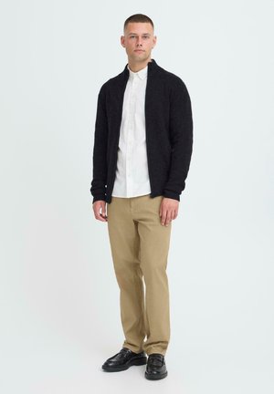 Black zip-up cardigan over a white button-down shirt, paired with tan pants and black shoes. The fabric appears textured and casual.