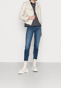 Hellebeige Leder-Motorradjacke, marinenblau-weiß gestreiftes Hemd, blaue enge Jeans, weiße Stiefeletten mit beigen Akzenten und gerippten Sohlen.