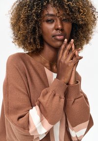 Vrouw met krullend haar, een bruine trui met witte strepen, vingers samengevouwen bij de kin, een klein kruisje als ketting draagend, neutrale achtergrond.