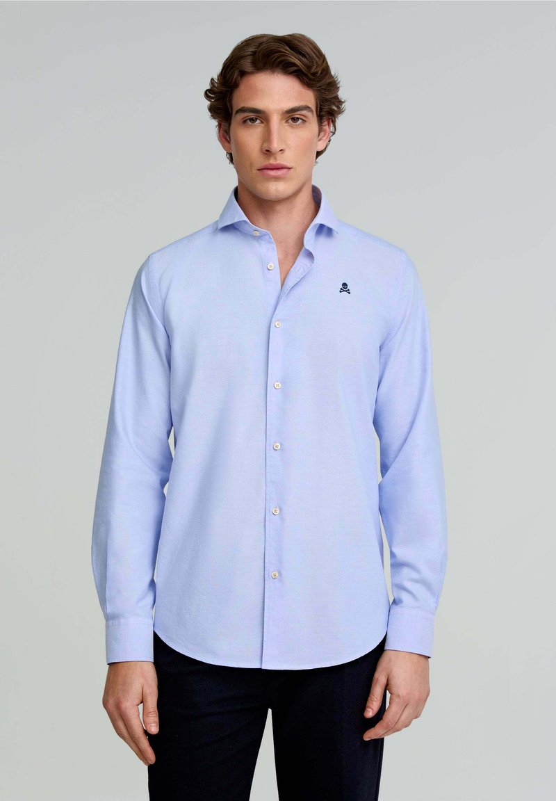 Camicia a maniche lunghe azzurro chiaro con colletto a bottoni, con un piccolo logo di teschio ricamato sul lato sinistro del petto e bottoni color crema.