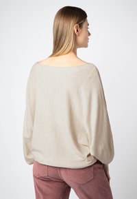 Beige oversized gebreide trui met een brede bootnek en lange mouwen, gecombineerd met roze corduroy broeken, die een textuurdetail hebben.