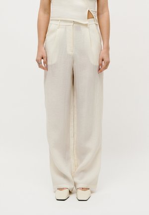 Nő magasderekú, bőszárú krémszínű nadrágot, hozzá illő ujjatlan felsőt és négyzet orrú krémszínű cipőt visel, egyszínű háttér előtt.