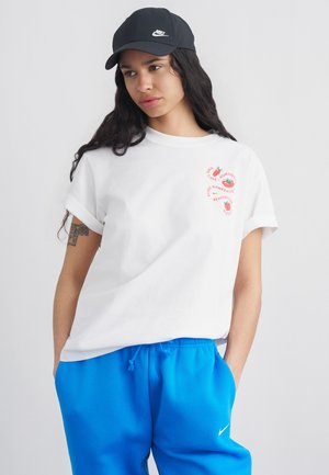 Młoda kobieta z długimi ciemnymi włosami, nosząca czarną czapkę Nike, białą koszulkę z czerwonymi grafikami oraz jaskrawo niebieskie spodnie dresowe Nike, dłonie w kieszeniach.