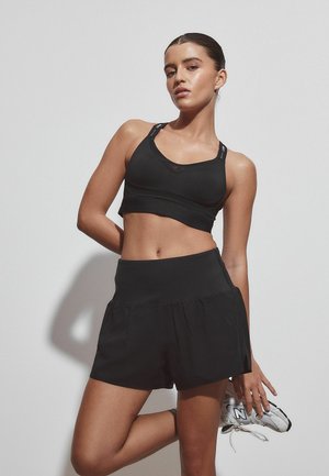 Kobieta w czarnym sportowym biustonoszu i szortach trzymająca białego trampka za plecami, pozująca na jasnym jednolitym tle.